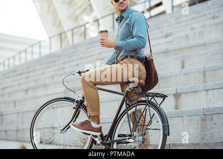 Ritagliato colpo di uomo in auricolari seduto sulla moto e tenendo monouso tazza di caffè sulla strada Foto Stock