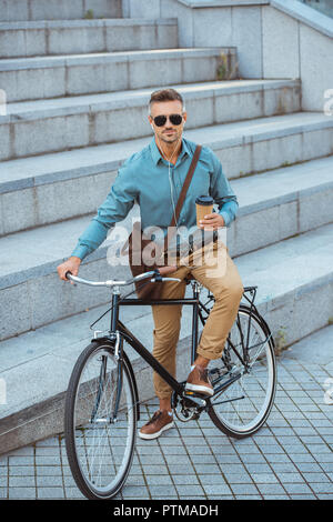 Uomo bello in occhiali da sole azienda Caffè di andare e guardando la fotocamera mentre è seduto in bici su strada Foto Stock