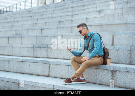 Bello elegante uomo in auricolari e occhiali da sole seduti sulle scale e utilizza lo smartphone Foto Stock