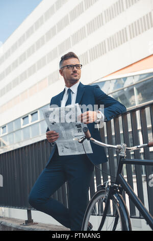 Persone di mezza età buisnessman in tuta e occhiali azienda giornale e guardando lontano mentre in piedi con la bici su strada Foto Stock
