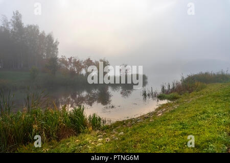 Tramonto su un tranquillo lago con riflessioni visto oltre a canne con la luce calda del sole e presto la nebbia Foto Stock