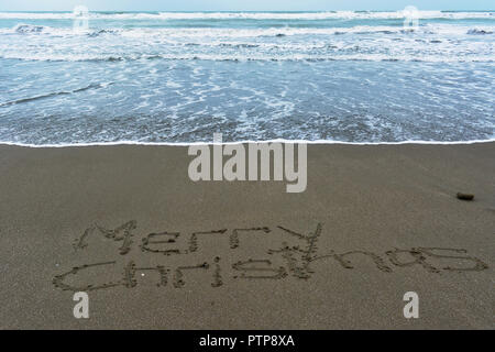 Buon Natale scritto a mano nella sabbia con surf dietro, creste bianche sulle piccole onde. Foto Stock