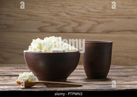 Cottage fresco formaggio nel recipiente di argilla con il cucchiaio di legno con un bicchiere di latte sul rustico sfondo di legno Foto Stock