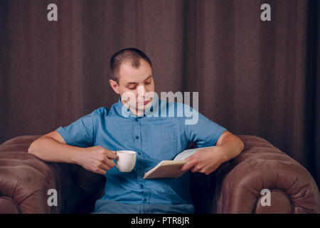 Uomo con un libro e una tazza di caffè nelle sue mani seduto in poltrona e la lettura. Psicologo. Psicoterapeuta. Foto Stock