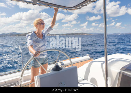 Attraente femmina bionda skipper navigazione la fantasia del catamarano a vela su soleggiate giornate estive sulla calma il mare blu dell'acqua. Foto Stock