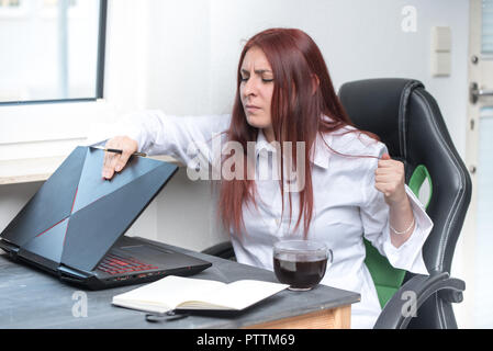 Un sottolineato, arrabbiato giovane donna è seduta alla sua scrivania e si sta chiudendo il suo computer portatile con una intensa rabbia Foto Stock