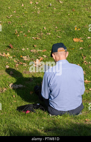 Un uomo seduto in un parco sull'erba la lettura di un libro e getta un' ombra indossando un estate cappello o berretto da baseball. Foto Stock