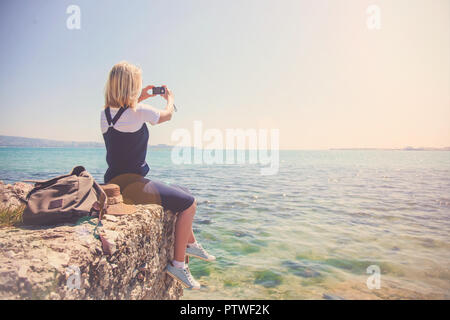 Ragazza giovane viaggiatore prende le foto sulla fotocamera bella vista dal mare. La ragazza ama al concetto di viaggio per i viaggiatori. Vista dal retro del per Foto Stock