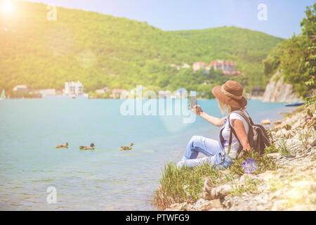 Ragazza traveler si siede su un lago di montagna e scatta foto ragazza prende la foto per il blog di viaggio. Vista dal retro del viaggiatore turistico Foto Stock