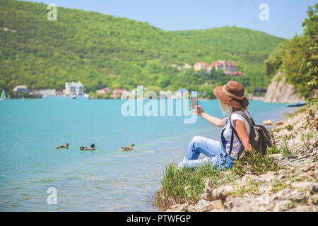 Ragazza traveler si siede su un lago di montagna e scatta foto ragazza prende la foto per il blog di viaggio. Vista dal retro del viaggiatore turistico Foto Stock