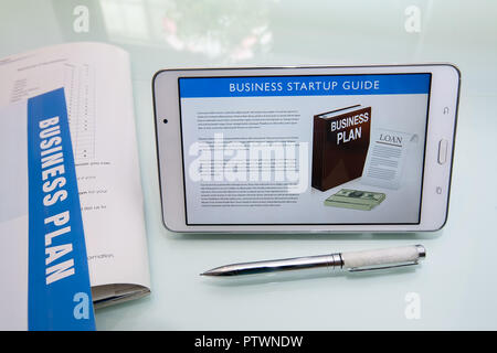 Scrivere un business plan utilizzando un business guida di avvio, materiale collaterale a stampa, penna e tavoletta o dispositivo mobile su un vetro riflettente desk. Foto Stock