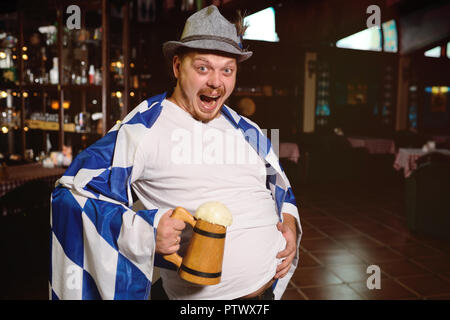 Allegro uomo grasso con un boccale di birra e con un oktoberfest flag in un cappello bavarese su un sfondo di pub Foto Stock