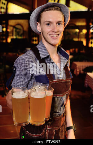 Un giovane uomo attraente in tradizionali abiti bavarese e un cappello con una piuma tiene nelle sue mani un sacco di bicchieri di birra sullo sfondo pub duri Foto Stock