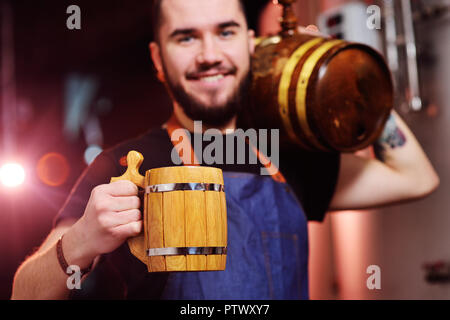 Giovani attraenti birraio barbuto con un boccale di legno nelle sue mani e con un barilotto di birra sorridente sullo sfondo della birreria Foto Stock