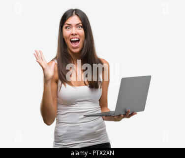 Giovane donna ispanica utilizzando computer portatile molto felici ed eccitati, vincitore espressione celebrando la vittoria urlando con un grande sorriso e le mani alzate Foto Stock