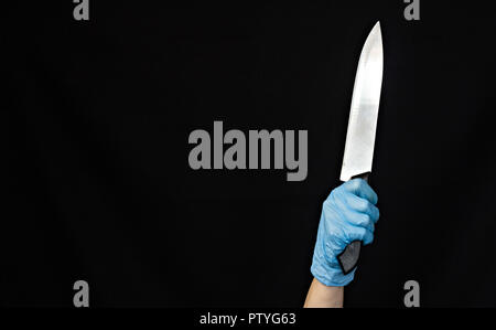 La mano di una ragazza in un blu guanto medicale con un coltello su un sfondo nero, close-up Foto Stock