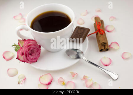 Caffè con bastoncini di cannella, rose, cioccolato fetta su uno sfondo bianco, petali di rosa Foto Stock