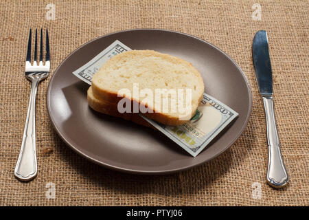 Due fette di pane con un centinaio di dollari americani bill come hamburger di denaro sulla piastra marrone con coltello e forchetta. Il denaro pronto per essere consumato. Concetto Foto Stock