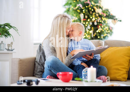 Una madre e un piccolo ragazzo seduto su un divano di casa al tempo di Natale, la lettura di un libro. Foto Stock