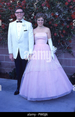 1962, America, andando a prom.....un giovane uomo che indossa uno smoking bianco e e una giovane donna che indossa una lunga frilly abito rosa di stare in piedi insieme a un'immagine prima di frequentare la PROM, un importante e tradizionale fine del termine di danza e un evento sociale per noi studenti di scuola superiore. Negli anni cinquanta e sessanta, PROM è stato più di un paio di evento, dove un ragazzo chiede ad una ragazza per essere la sua data e alla scorta di lei dopo il prelievo di lei fino a casa sua e con un sacco di foto scattata! Foto Stock