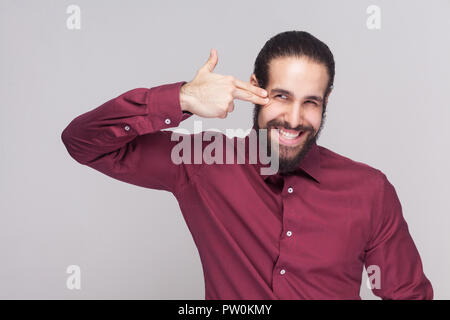 Ritratto di crazy uomo bello con scuri raccolti i capelli lunghi e la barba in maglietta rossa in piedi e tenere la mano sulla testa con la pistola gesto. piscina studio Foto Stock