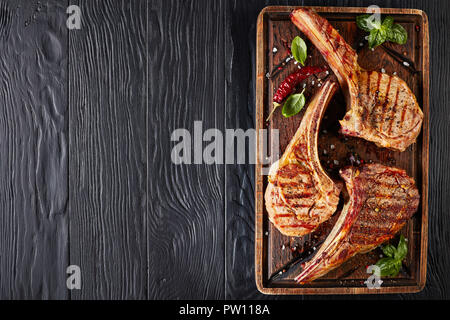 Tre età secco grigliate di tomahawk bistecche o cowboy bistecche di manzo su un vecchio rude tagliere di legno con peperoncino essiccato, il sale e le foglie di basilico, close-up, fl Foto Stock