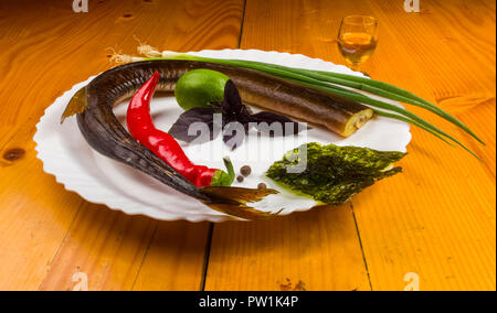 Still Life - aguglia affumicato con calce, basilico, cipollotti, peperoncino, nori chips, spezie, olio d'oliva in un bianco piatto di ceramica, su di un tavolo di legno Foto Stock