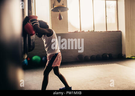 Il pugilato kid appoggiata contro un sacco da boxe indossando guanti da boxe. Stanco boxing kid in piedi in un boxing palestra. Foto Stock