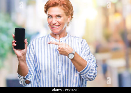 Accattivante senior redhead caucasica donna che mostra lo schermo dello smartphone su sfondo isolato molto felice puntando con mani e dita Foto Stock