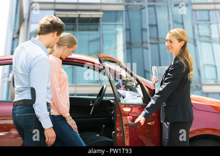 Donna sorridente apertura del Concessionario Auto Rossa porta per Coppia Giovane Foto Stock