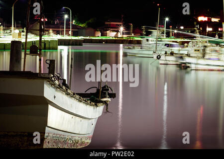 Un vecchio, piccole e bianche barca da pesca parcheggiato di notte sulla riva. Una lunga esposizione shot e acqua è di colore violaceo con la riflessione della luce. Foto Stock