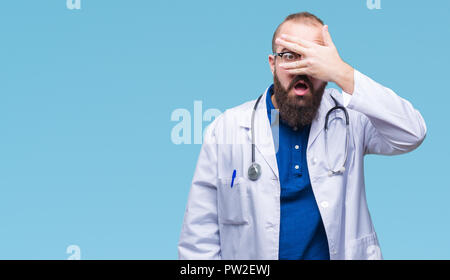 Giovane medico caucasica uomo che indossa medical mantello bianco su sfondo isolato peeking in stato di shock che copre il viso e gli occhi con la mano, guardando attraverso fing Foto Stock