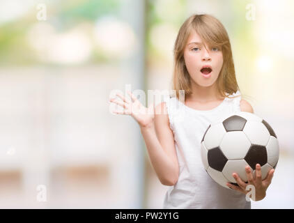 Giovane biondo toddler tenendo palla calcio molto felici ed eccitati, vincitore espressione celebrando la vittoria urlando con un grande sorriso e le mani alzate Foto Stock