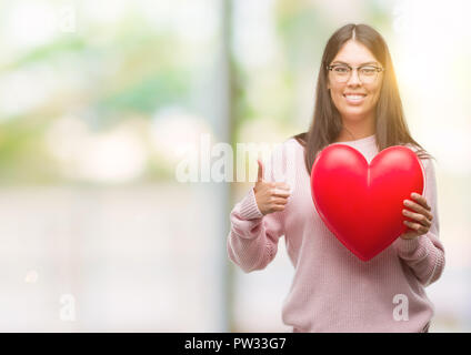 Giovane donna ispanica tenendo il cuore in amore felice con un grande sorriso facendo segno ok, pollice in alto con le dita, segno eccellente Foto Stock