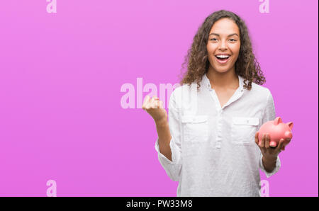 Giovane donna ispanica holding salvadanaio urlando orgoglioso e celebrando la vittoria e il successo molto eccitato, allietarla emozione Foto Stock