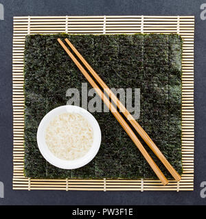 Sushi ingredienti, nori alghe foglio, materie di riso sushi e bacchette in bambù makisu mat, fotografato overhead su ardesia Foto Stock
