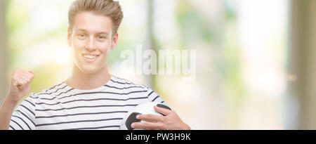 Giovane bella bionda uomo tenendo palla calcio urlando orgoglioso e celebrando la vittoria e il successo molto eccitato, allietarla emozione Foto Stock