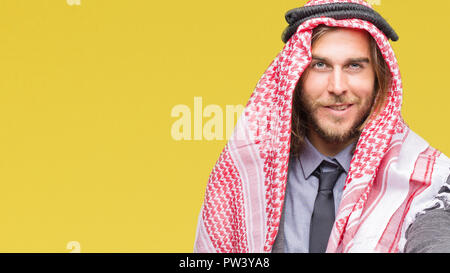 Giovane bello arabian uomo con capelli lunghi che indossa keffiyeh su sfondo isolato sorridente cordiale offrendo stretta di mano come saluto e accogliente. Suc Foto Stock