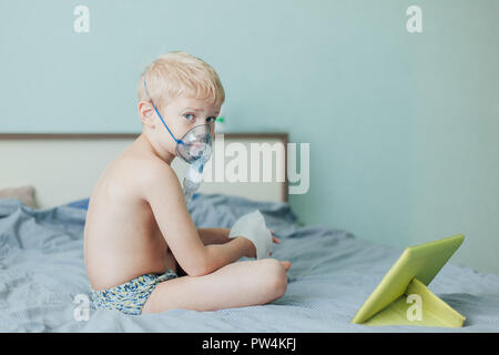 Piccolo Ragazzo non inalazione terapeutico usando un nebulizzatore e guarda i cartoni sulla tavoletta Foto Stock