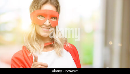 Giovane super eroe donna che indossa il capo sorridente largamente che mostra i pollici fino al gesto fotocamera, espressione di simili e approvazione Foto Stock