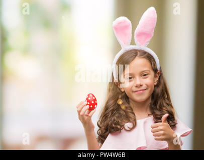 Bruna ragazza ispanica che indossa la pasqua orecchie di coniglio felice con un grande sorriso facendo segno ok, pollice in alto con le dita, segno eccellente Foto Stock