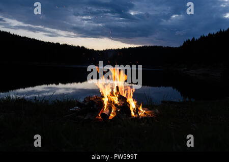 Campfire dopo il tramonto in montagna accanto a un lago Foto Stock