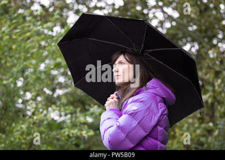 Vista laterale della donna che indossa un cappotto isolato all'aperto che tiene un ombrello nero e guarda verso l'alto. Foto Stock
