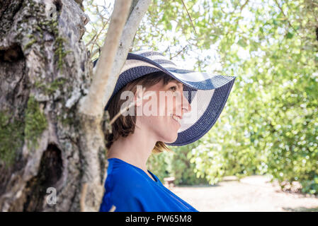 Donna attraente in una vasta colmato cappello in piedi appoggiata contro un albero in ombra in un caldo giorno d'estate e di sole. Foto Stock