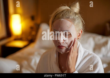 La donna è l'applicazione di fogli di maschera sul viso in camera da letto Foto Stock