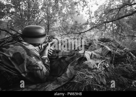 Unidentified ri-enactor vestito come il tedesco della Wehrmacht Soldato di fanteria nella guerra mondiale II seduta nascosti con un fucile arma in un agguato in trincea in autunno Foto Stock