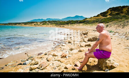 Uomo di mezza età seduto da solo su una spiaggia rocciosa, guardando verso il mare Mediterraneo, contemplando la vita, nord, Cipro. Foto Stock