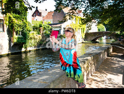 Donna seduta da sola su un muro basso a riva del canale, prendendo selfie di sé e la vista panoramica, città di Brugge, Belgio. Foto Stock