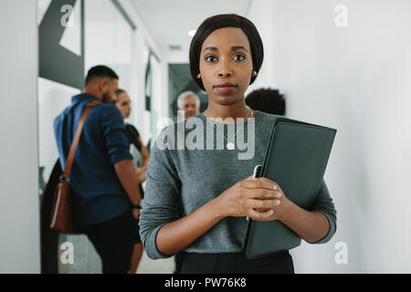 Fiducioso donna africana tenendo un file office corridoio con i suoi colleghi parlano in background. Executive femmina in piedi di fronte con business t Foto Stock