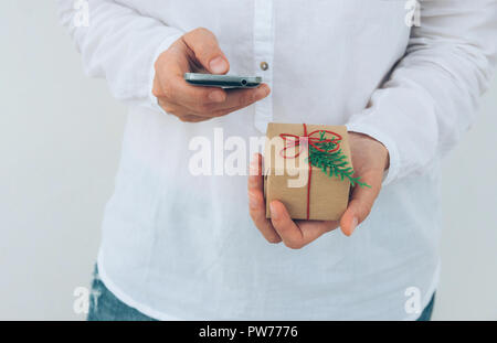Giovane donna caucasica in jeans shirt detiene in mani confezione regalo avvolto in marrone carta craft legata con lo spago e lo smartphone. Muro Bianco sullo sfondo. Chris Foto Stock